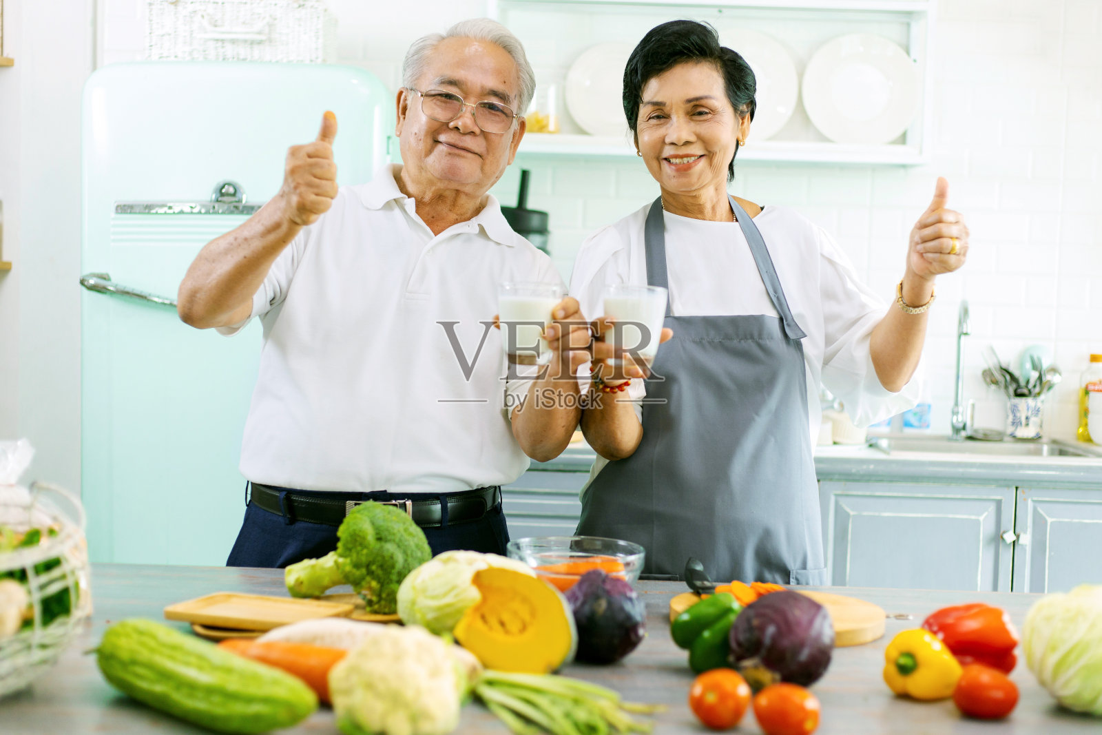 一个年长的亚洲夫妇在家庭厨房做饭的肖像。他们有一个微笑的脸，快乐，并显示他们的牛奶在他们的手中。退休人员喝牛奶使骨骼强壮。预防骨质疏松症。照片摄影图片