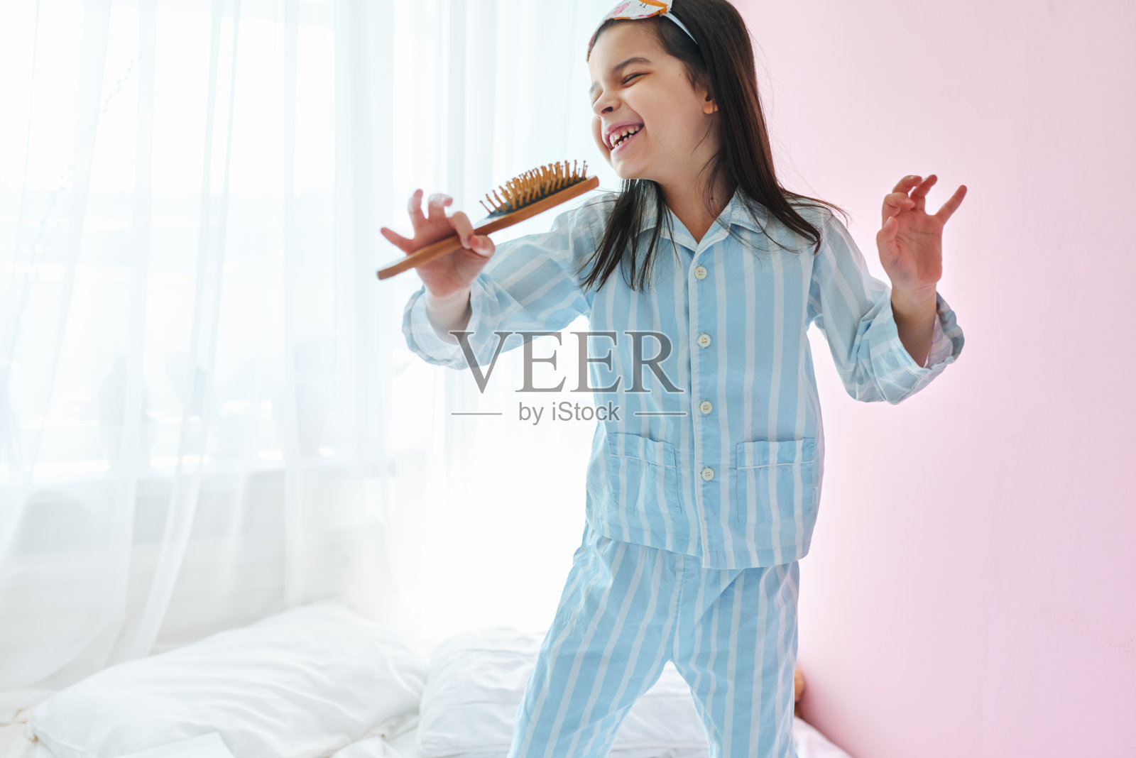 好玩的小女孩穿着睡衣在床上拿着梳子像麦克风唱歌模仿自己一个真正的歌手早上。照片摄影图片