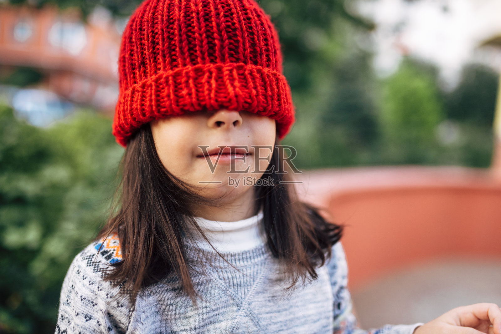 一个可爱的小女孩的肖像，戴着红色的针织帽，遮住了她的眼睛休息期间放学后。一个漂亮的孩子在户外玩得很开心。照片摄影图片
