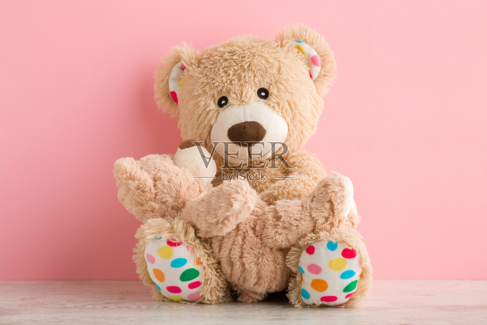 棕色的泰迪熊妈妈坐在和抱着她的孩子在浅粉色的墙壁背景桌上。可爱的、情感的时刻。特写镜头。前视图。照片摄影图片