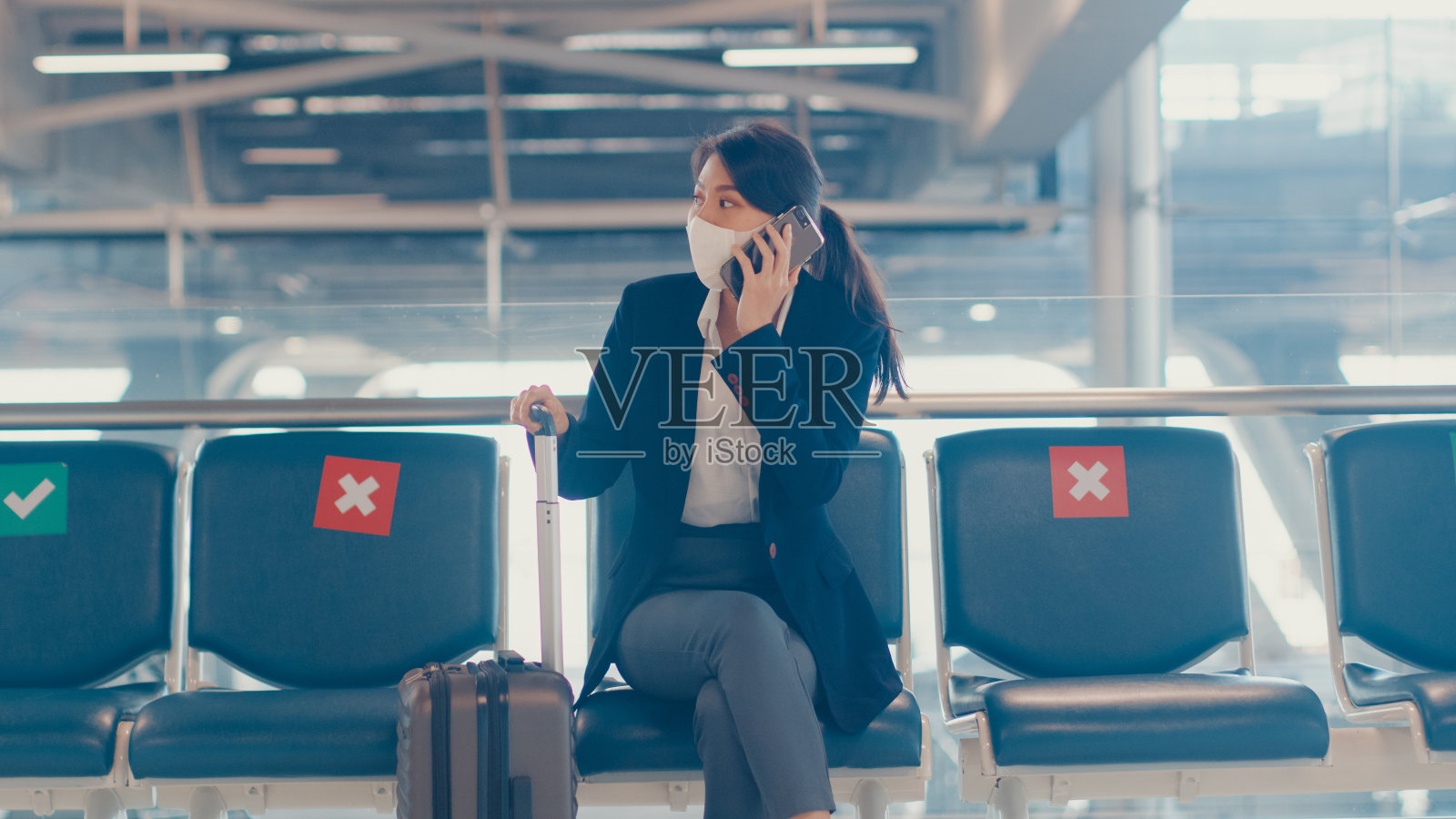 聪明的亚洲商务女孩穿着西装坐在手提箱在长凳打电话智能电话与伙伴在机场等待航班。照片摄影图片