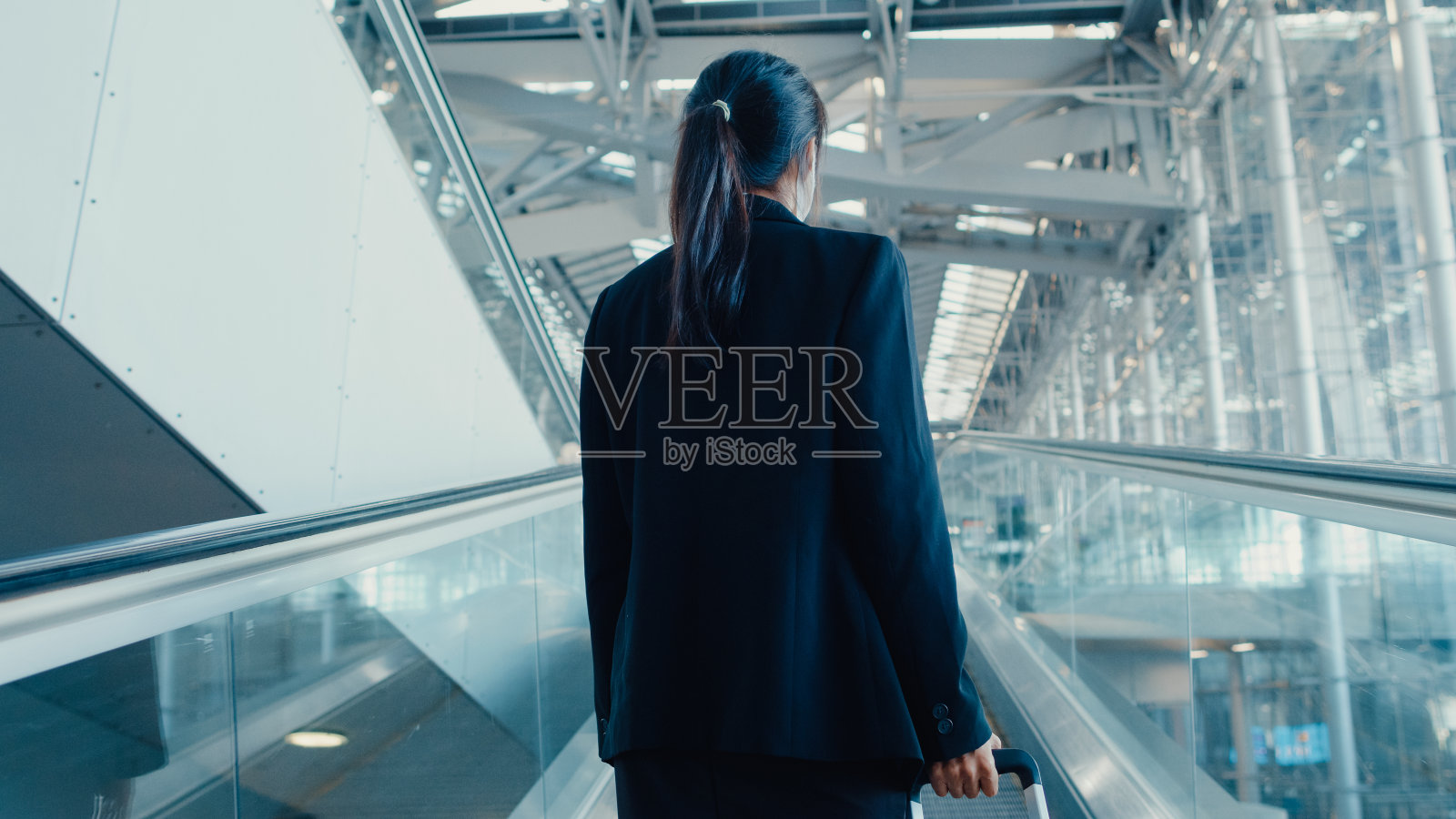 亚洲商务女孩戴着口罩拖着行李站在自动扶梯上四处张望步行到国际机场的终点站。照片摄影图片