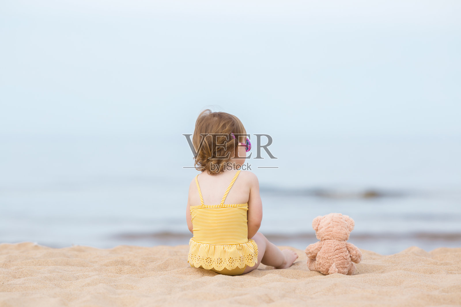 穿着黄色泳衣和棕色泰迪熊的小女孩坐在沙滩上，盯着温暖阳光明媚的夏日里的海水。后视图。看远。照片摄影图片
