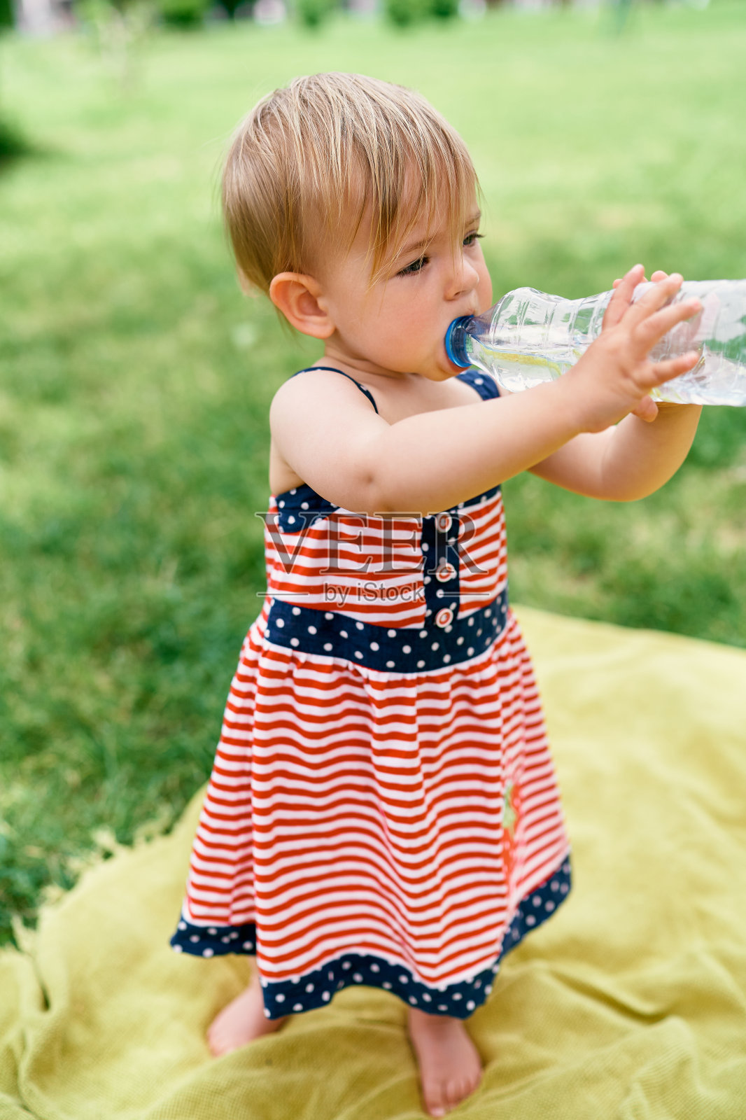 小女孩站在草坪上的毯子上从瓶子里喝水照片摄影图片