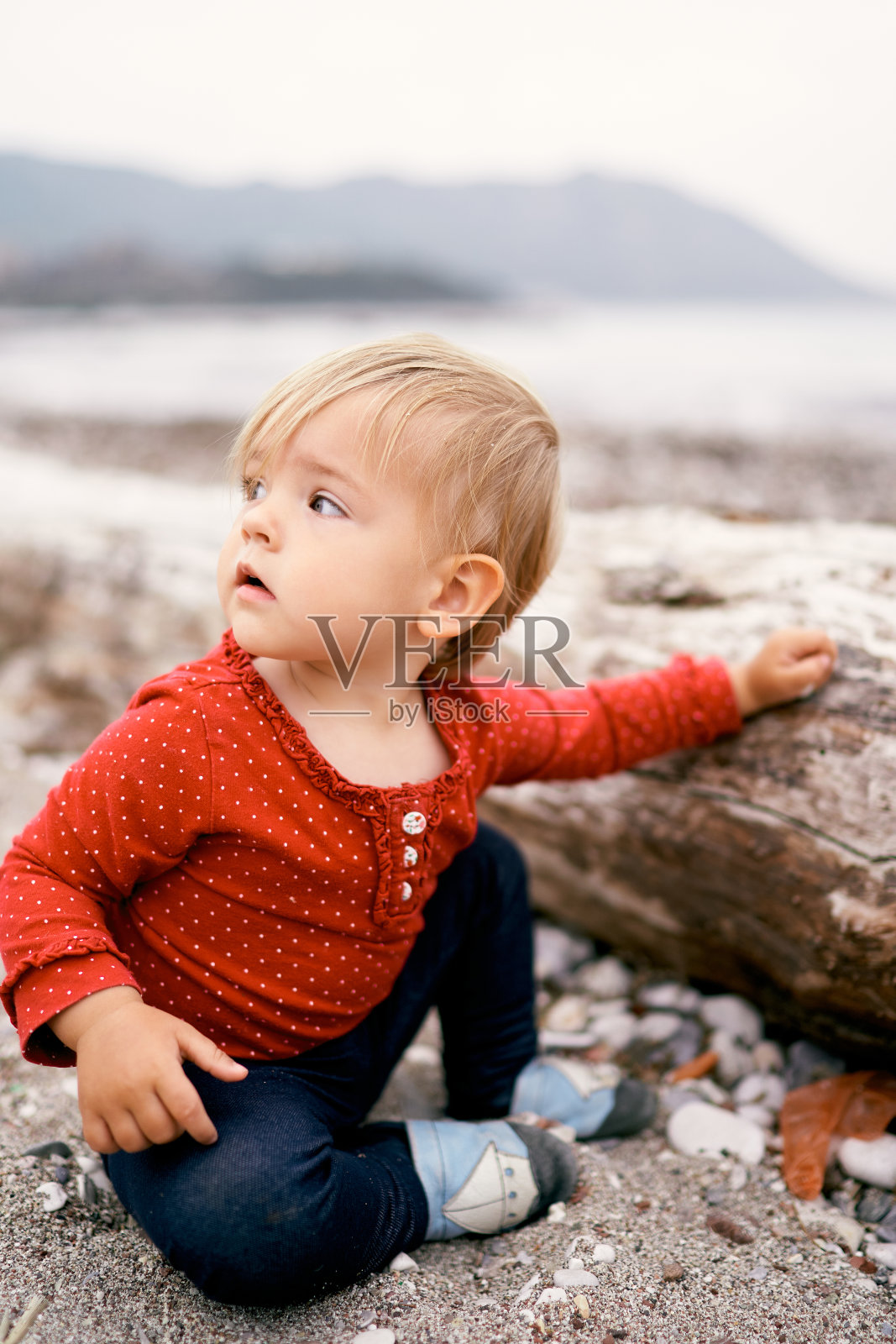 婴儿坐在一个旧浮木附近的卵石海滩上，把头转向一边照片摄影图片