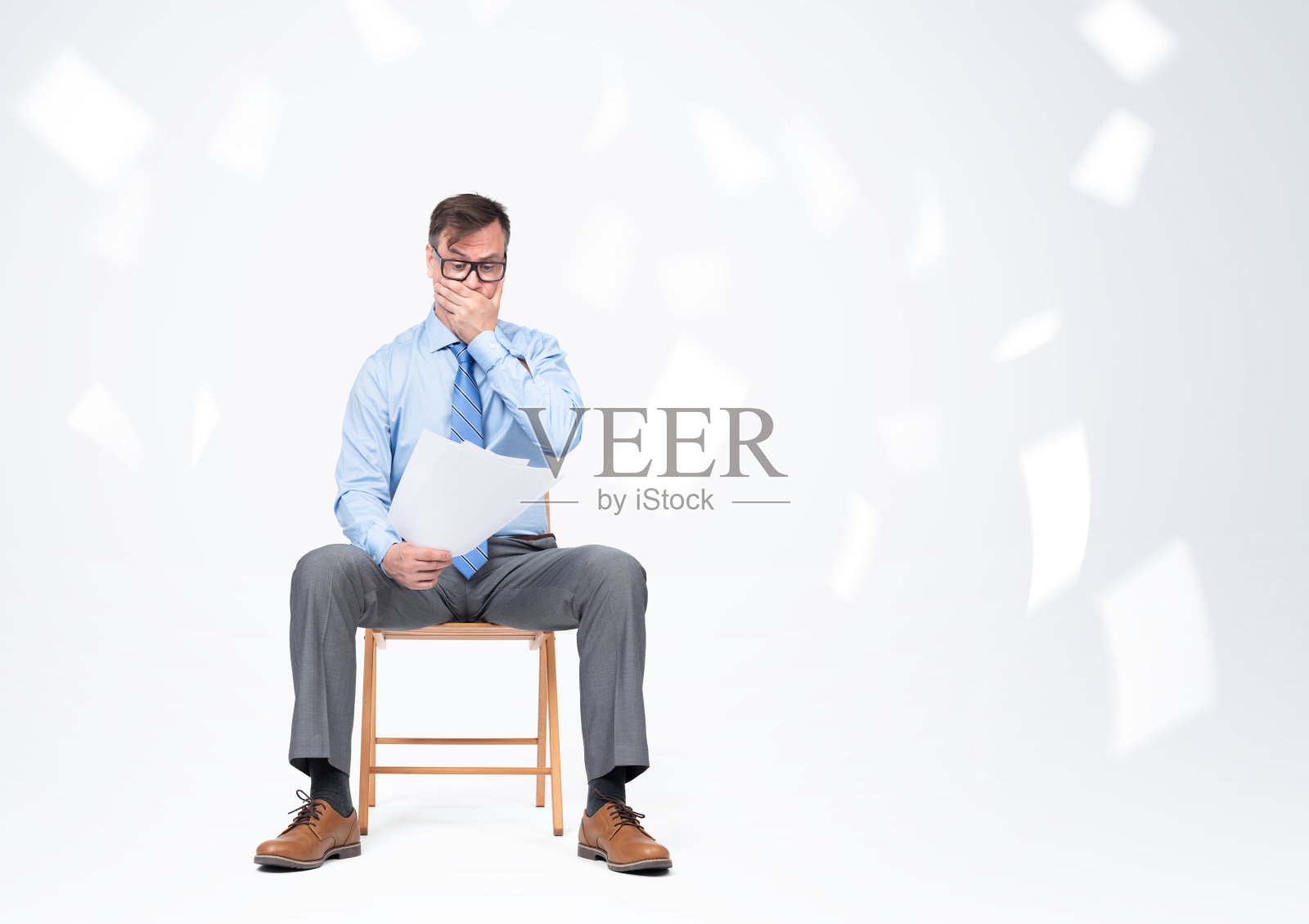 一个戴眼镜、穿衬衫打领带的商人坐在椅子上，手里拿着纸，背景很亮。照片摄影图片