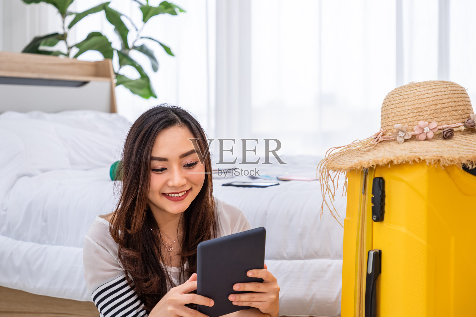 快乐的年轻亚洲女人坐在行李，准备行李箱暑假旅行，使用平板电脑计划和购买机票。照片摄影图片