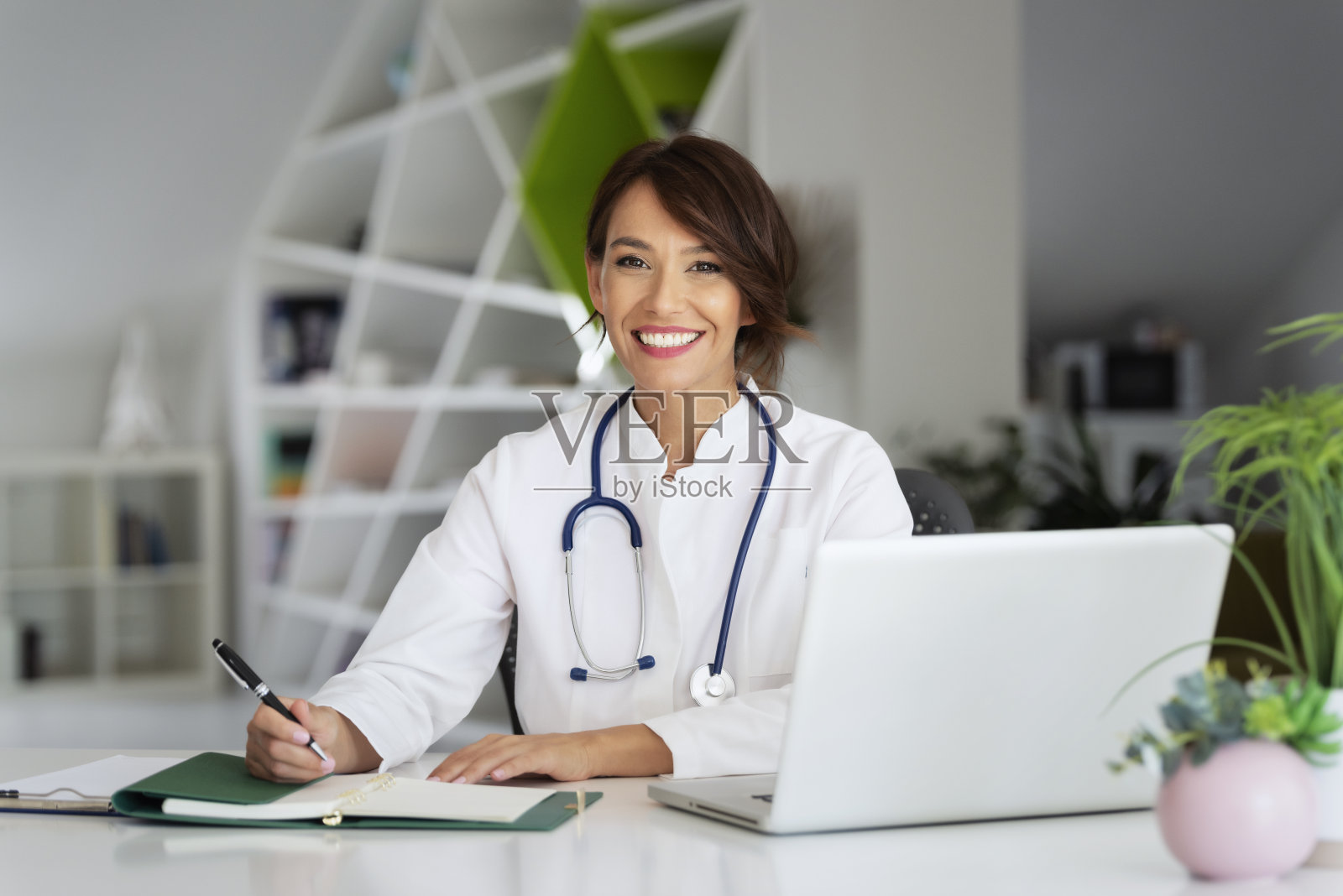 微笑的女医生使用笔记本电脑和写东西，而坐在医生的办公室照片摄影图片