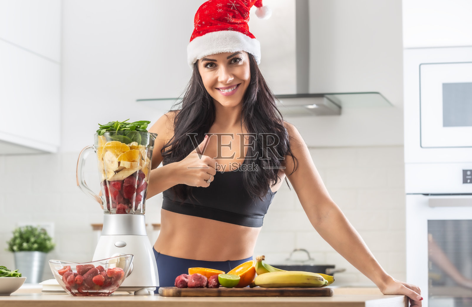 一个戴着圣诞帽、穿着运动服站在装满健康食品的健康奶昔机旁边的女人竖起大拇指。照片摄影图片