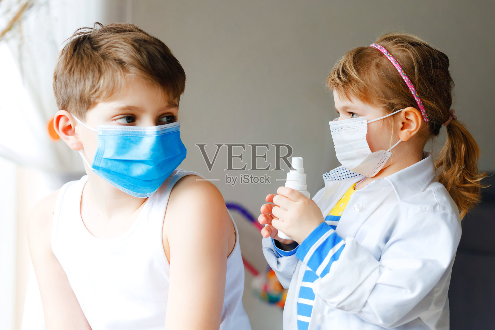 小女孩给弟弟打针，学弟学弟。孩子，兄弟姐妹带着医用口罩玩医生，拿着注射器注射疫苗。新冠病毒疫苗接种概念。孩子们玩角色游戏照片摄影图片