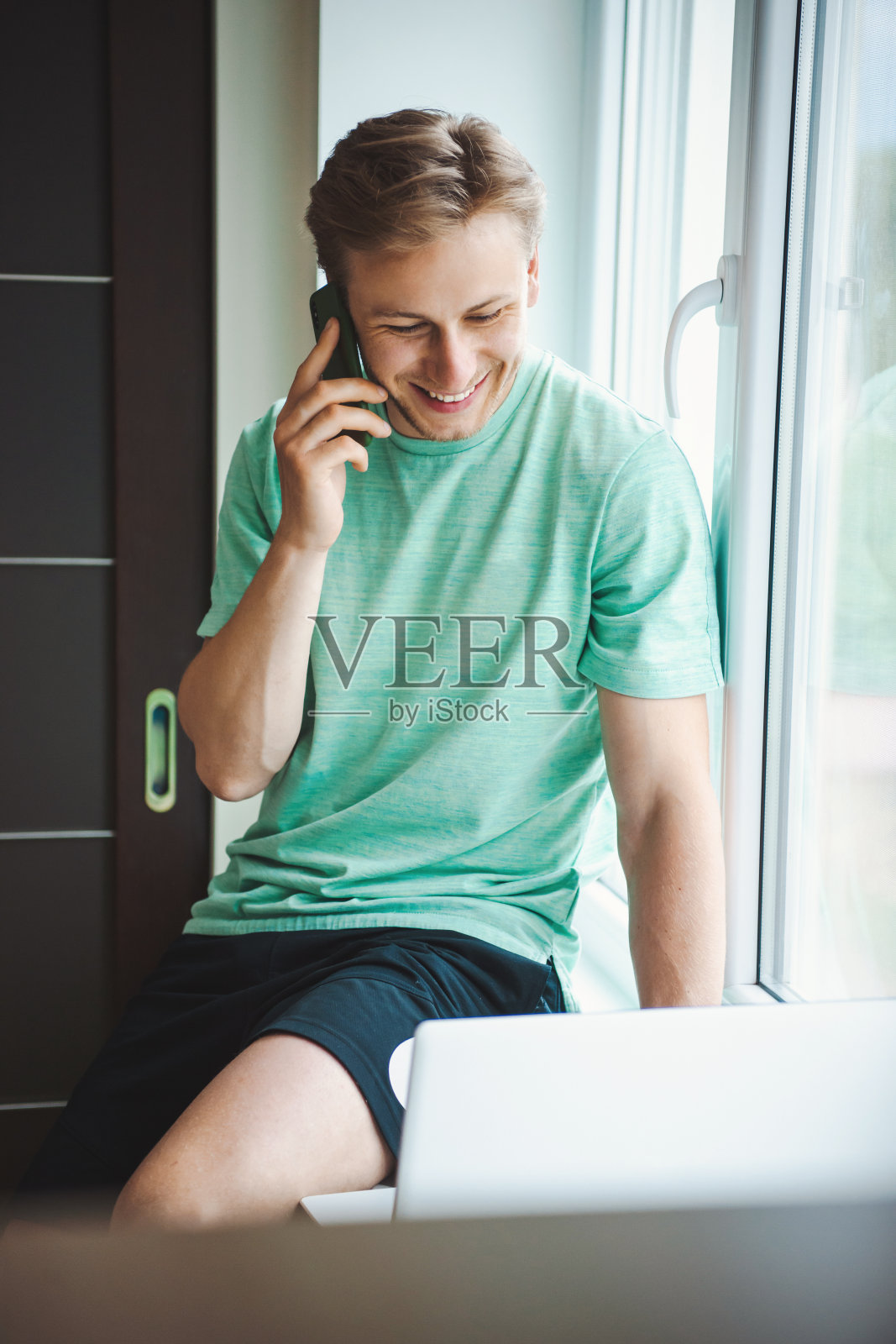 一个人坐在窗台上一边打电话一边在电脑上打字。在家工作。微笑的男人照片摄影图片