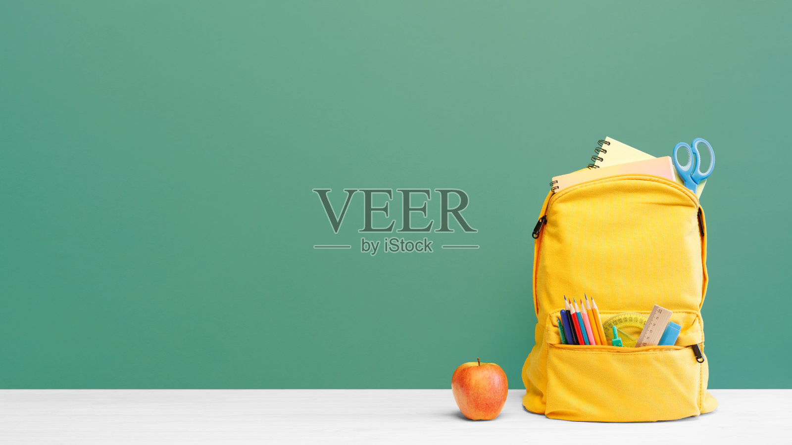双肩包不同的彩色文具和苹果在桌子上和绿色的背景。横幅设计回归学校概念背景。照片摄影图片