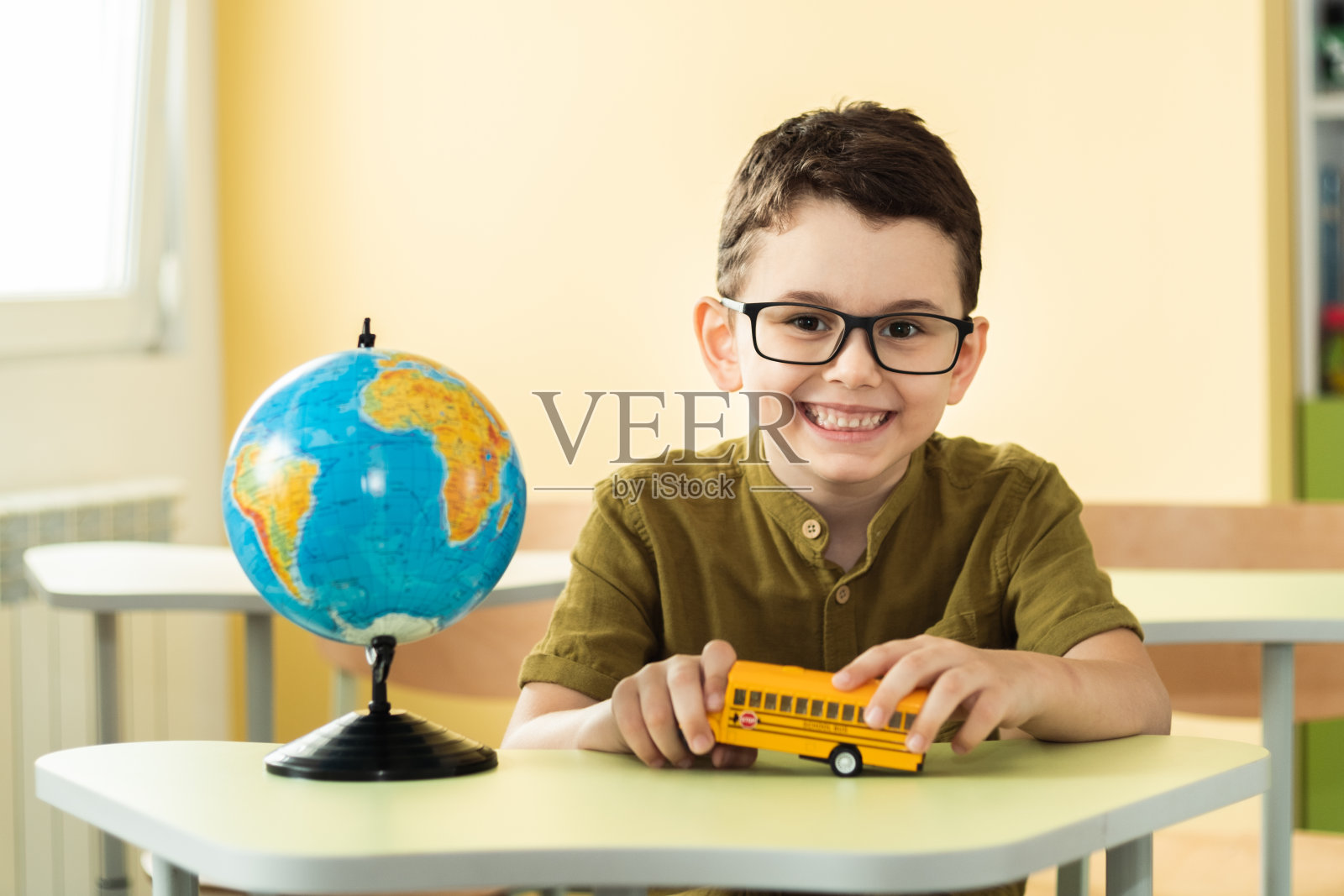 小学教室里戴着眼镜坐在书桌前的可爱白人男生。小男孩在地球仪旁边玩黄色校车玩具。回归学校理念照片摄影图片